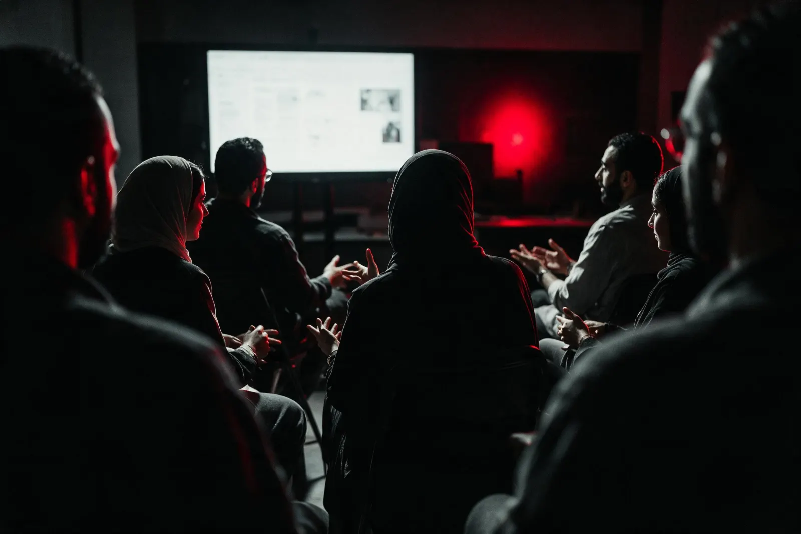 A live session in a dark room, a group seated facing a lit screen, mid-discussion.