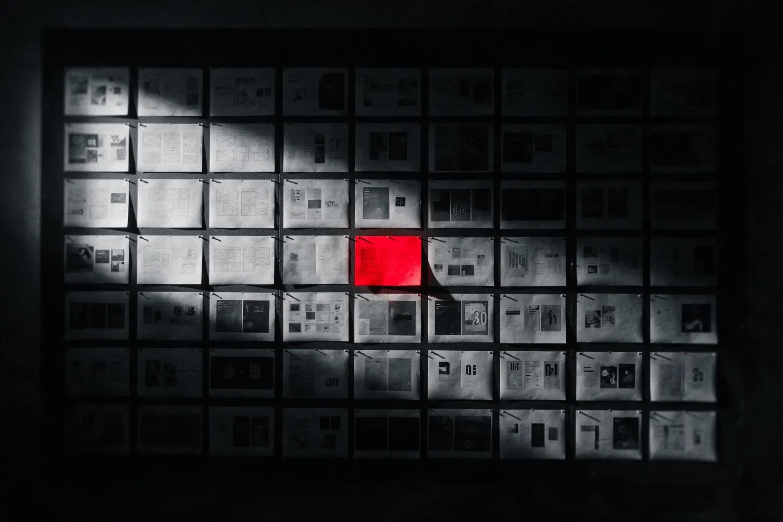 A wall of printed work pinned in a grid, with one sheet lit red.